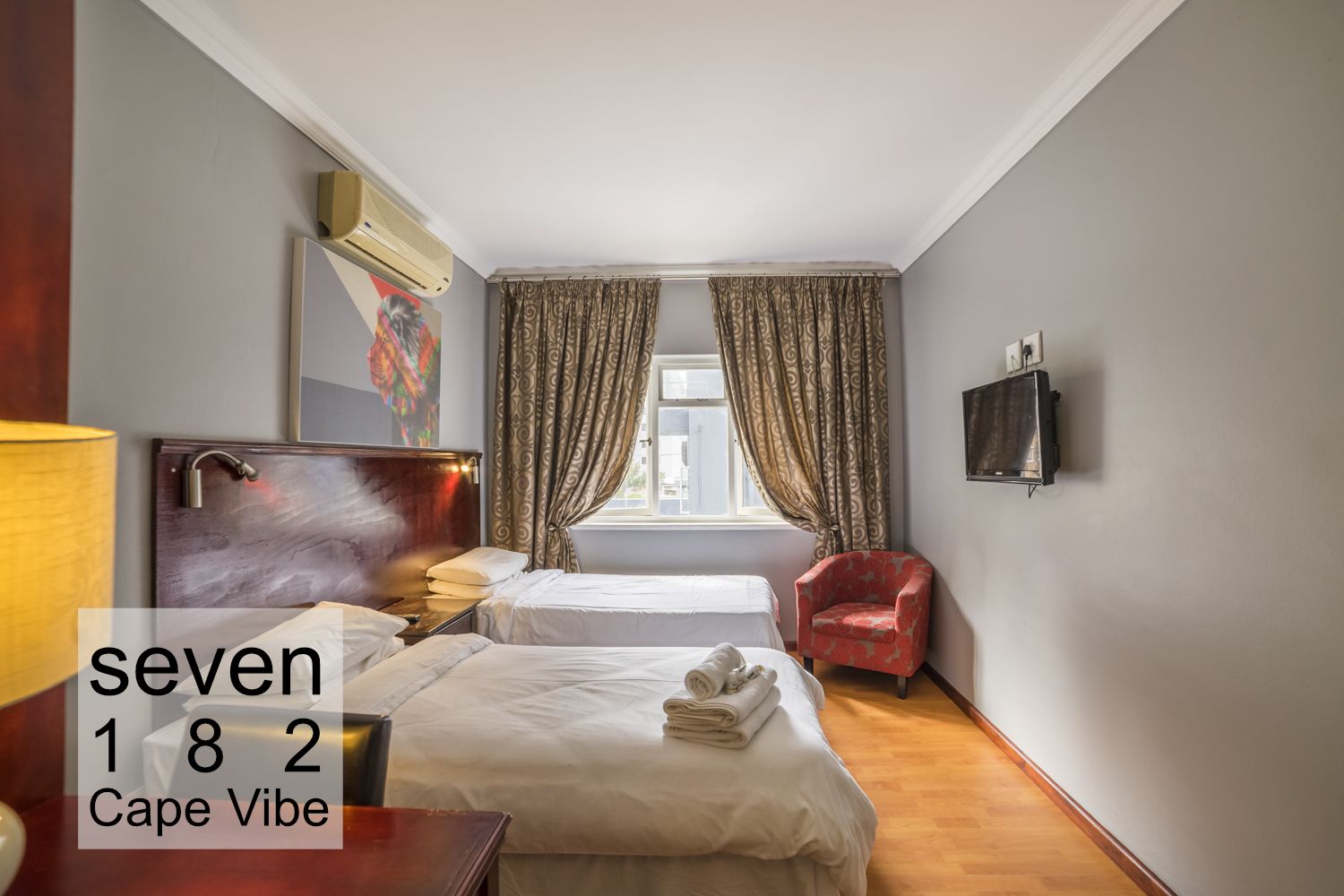 Una habitación de hotel con dos camas, un sillón rojo, un televisor y una ventana con cortinas.