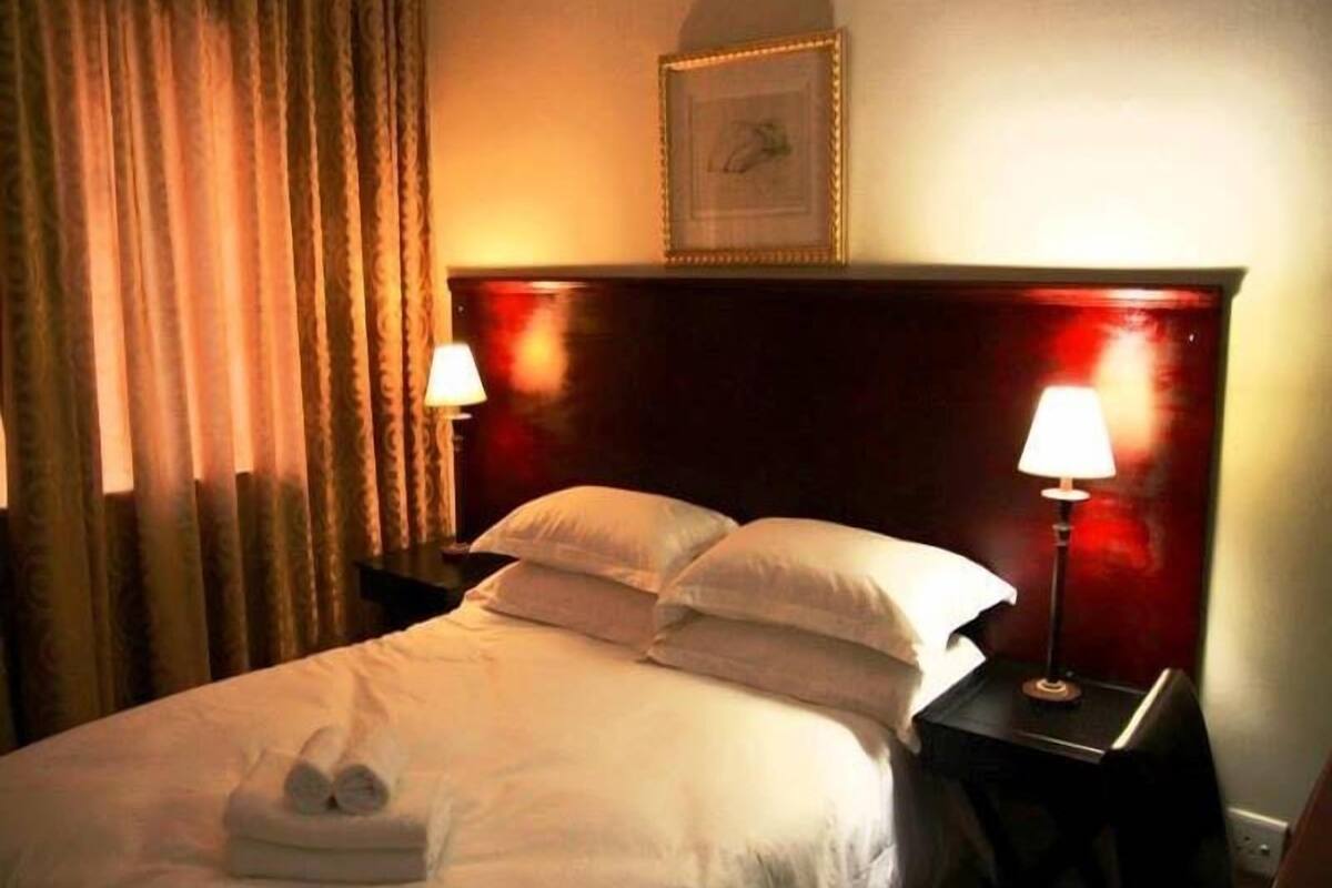 Une chambre d’hôtel avec un lit, deux lampes de chevet, une tête de lit, un tableau encadré au mur et une fenêtre avec des rideaux.