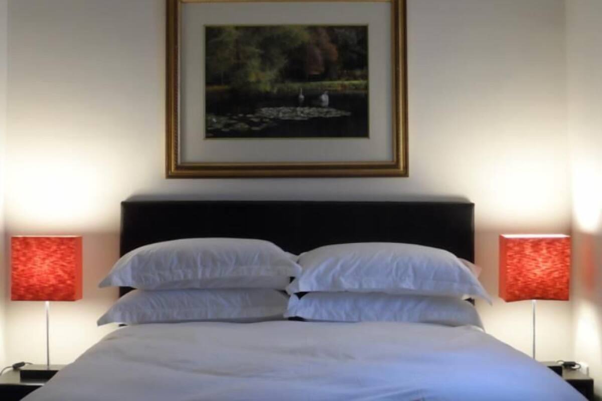 Un lit bien fait, avec du linge de lit blanc, une tête de lit sombre et deux lampes de chevet dotées d’abat-jours rouges.