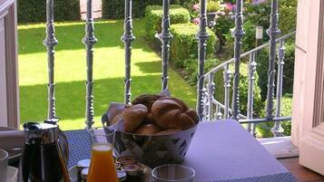 Een balkon met een metalen hekwerk, een tafel met een broodmand, een koffiezetapparaat en een glas sinaasappelsap.