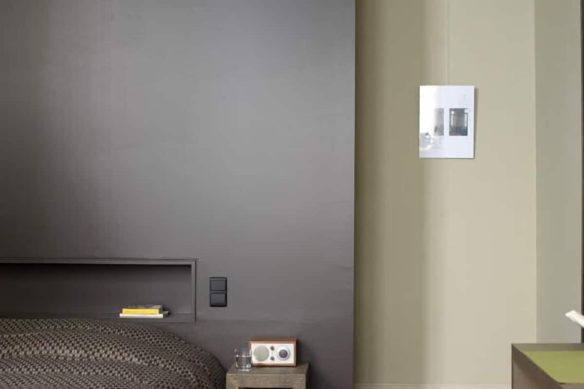 A room with a dark grey wall, a bed with a headboard, a bedside table with a radio, a small picture frame, and a bowl with fruits on a table.