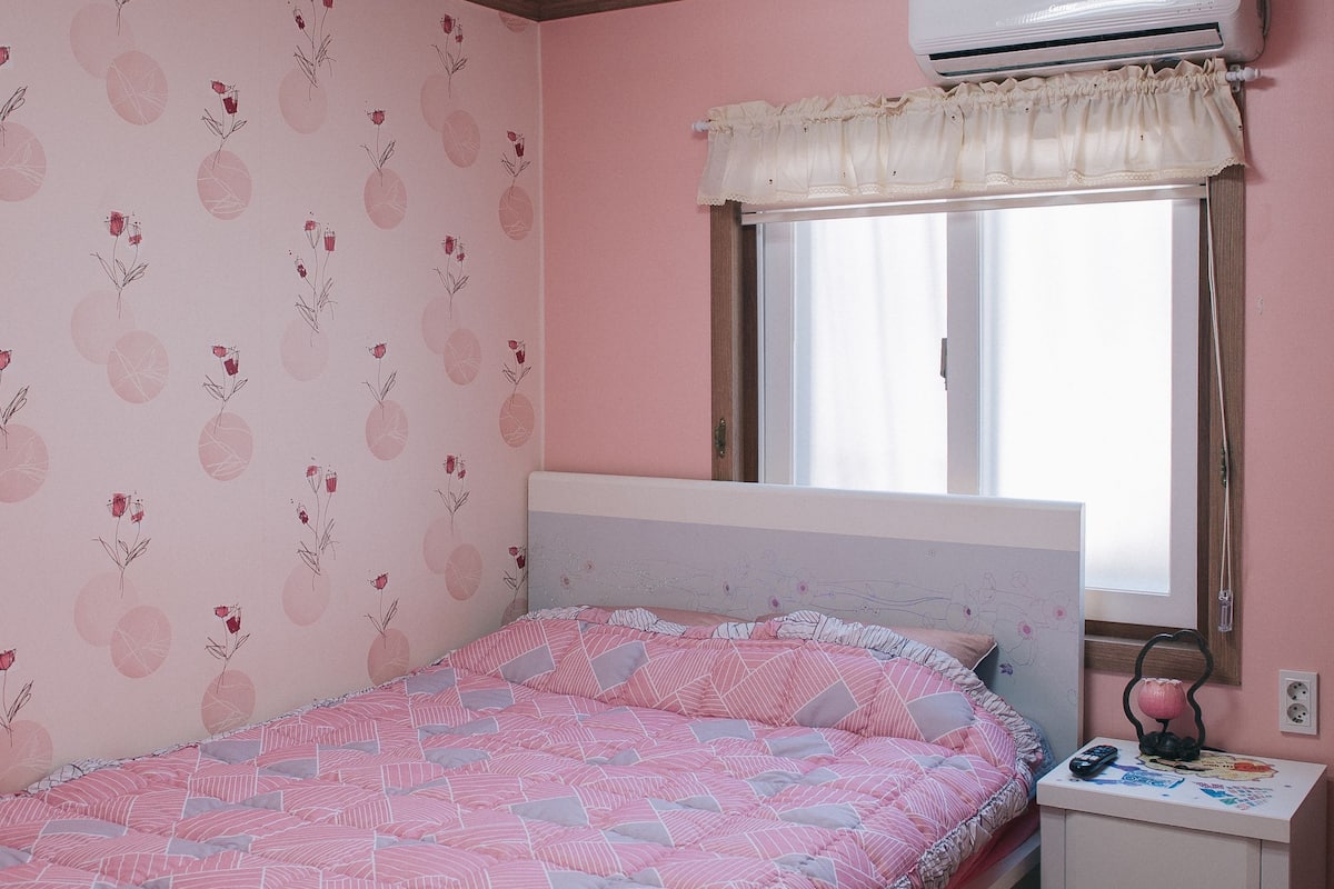 A bedroom with a bed, a nightstand, a window with a curtain, and an air conditioning unit.