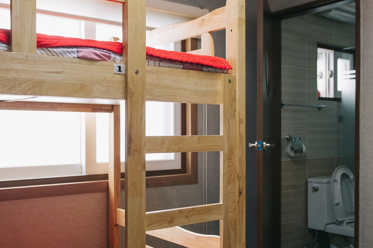 A bunk bed with red bedding, a wooden ladder, and a window. There is an air conditioning unit above the bed and a bathroom visible through an open door.