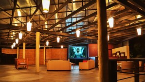 An indoor space with a high ceiling, wooden beams, and a large screen displaying a person. There are white sofas and a wooden chair. - Ranna 212 (Kahandamodara)