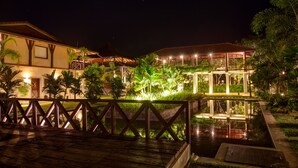 A nighttime view of a resort with a wooden deck, a pool, and well-lit buildings surrounded by greenery. - Ranna 212 (Kahandamodara)