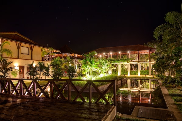 A nighttime view of a resort with a wooden deck, a pool, and well-lit buildings surrounded by greenery.