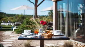 A table set with breakfast items, including a basket of pastries, a glass of orange juice, and cups, in an outdoor setting with a view of a garden and lounge chairs. - Relais des Vigiers (Monestier)