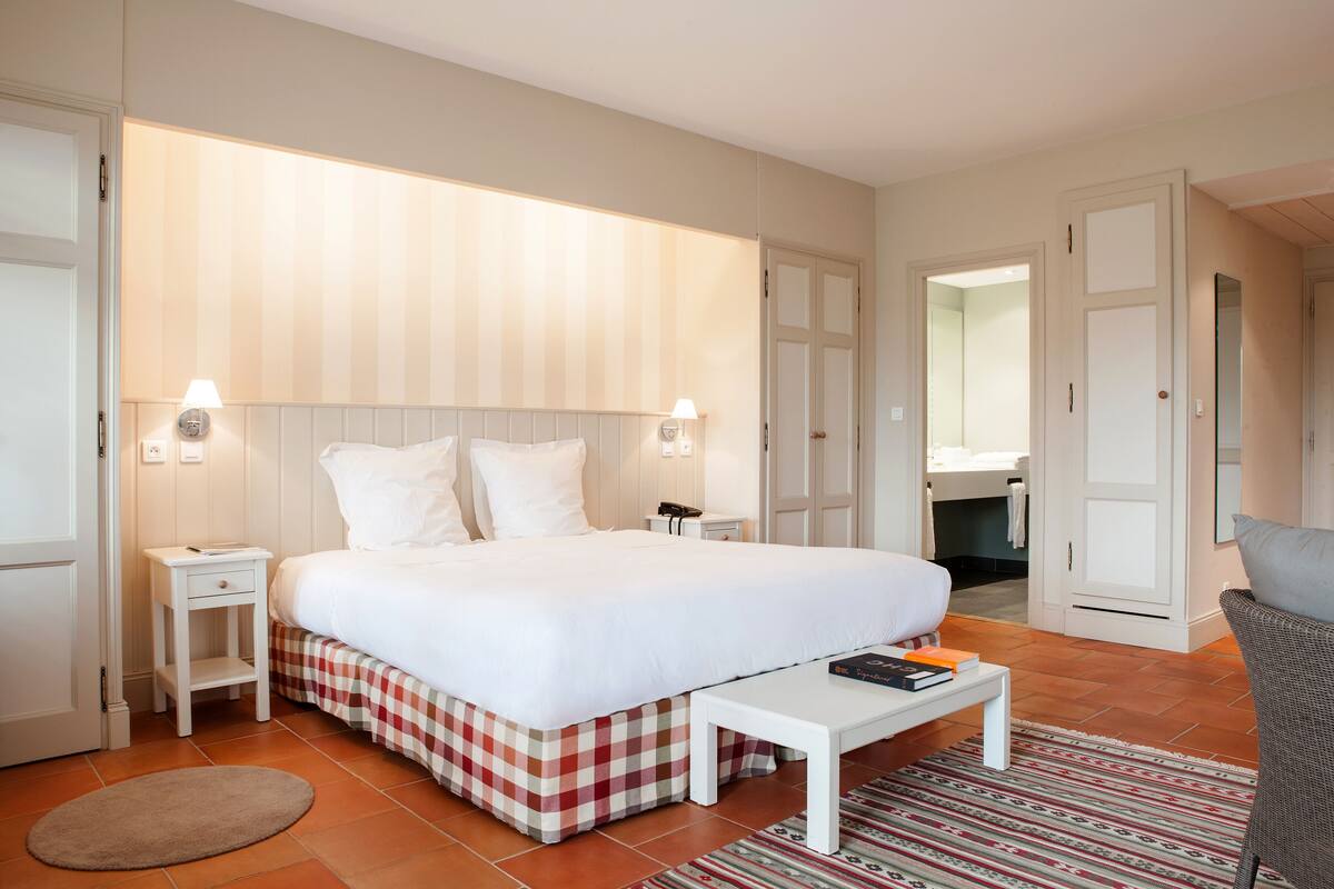 A hotel room with a large bed, bedside tables, a small table, a chair, a bathroom visible through an open door, and a patterned rug.