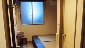 A narrow hallway with a wooden floor, a bed with a mattress and pillows, a small table with a chair, a window with blinds, and a door with a handle.