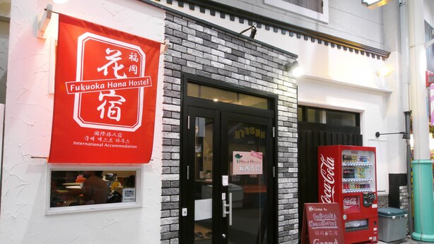 A Fukuoka Hana Hostel with a red sign, a black door, and a Coca-Cola vending machine outside.
