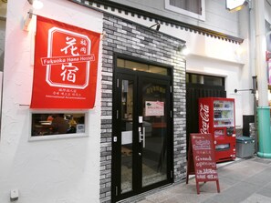 A Fukuoka Hana Hostel with a red sign, a black door, and a Coca-Cola vending machine outside.