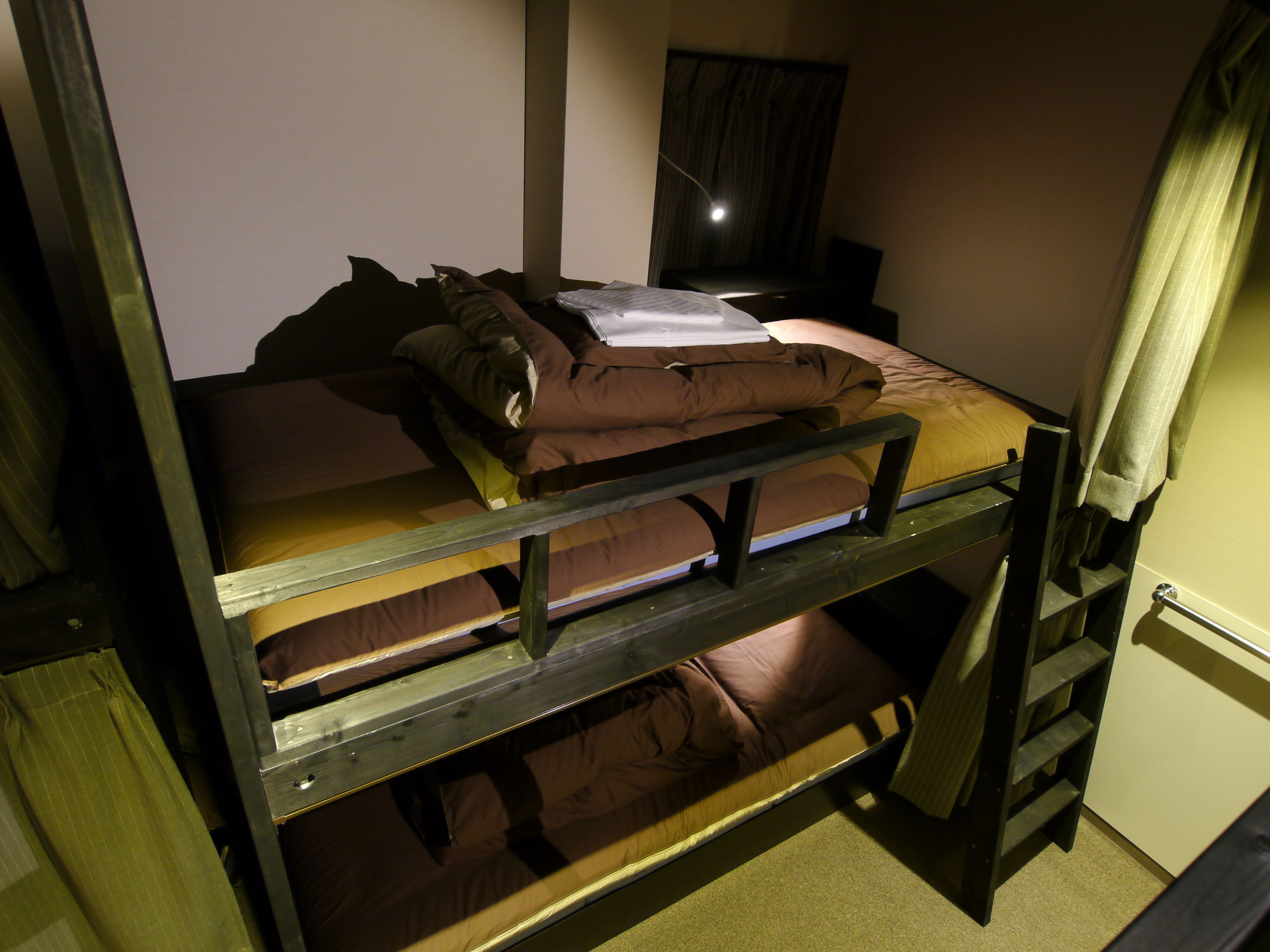 A bunk bed with a wooden frame, a mattress, and pillows. There is a nightstand with a lamp and a curtain.
