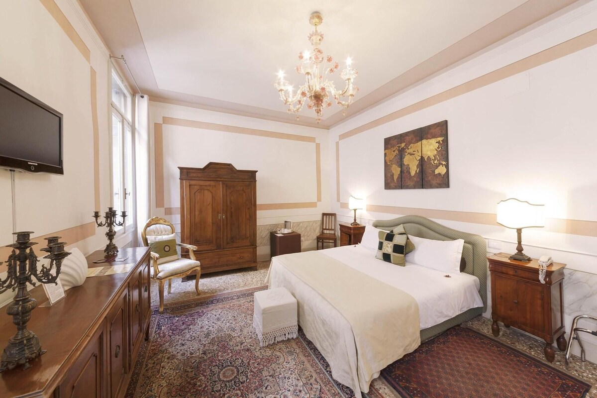 A hotel room with a large bed, a wooden wardrobe, a television, a chandelier, and a world map on the wall.