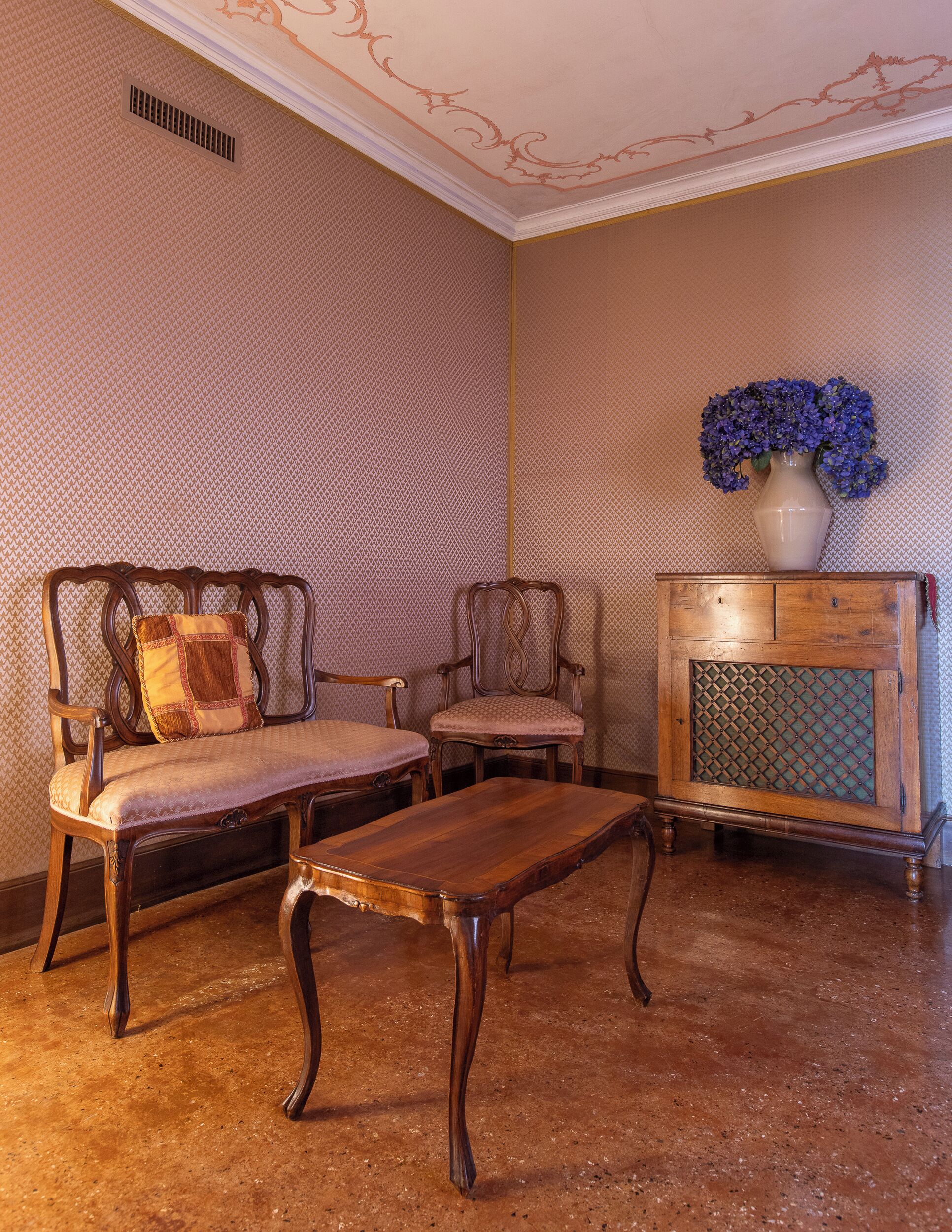 A room with a patterned wall, a wooden bench, two upholstered chairs, a small table, and a decorative vase with flowers.