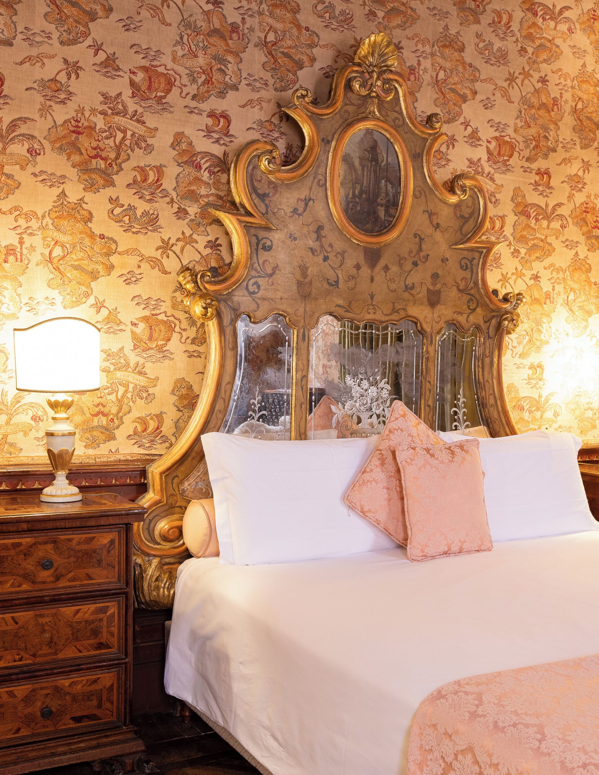 Un dormitorio con una cama grande y decorativa, una mesita de noche con lámpara y un espejo.