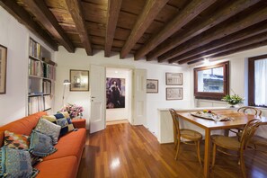 A cozy living room with a wooden ceiling, a dining table with chairs, a bookshelf, and a door leading to another room.