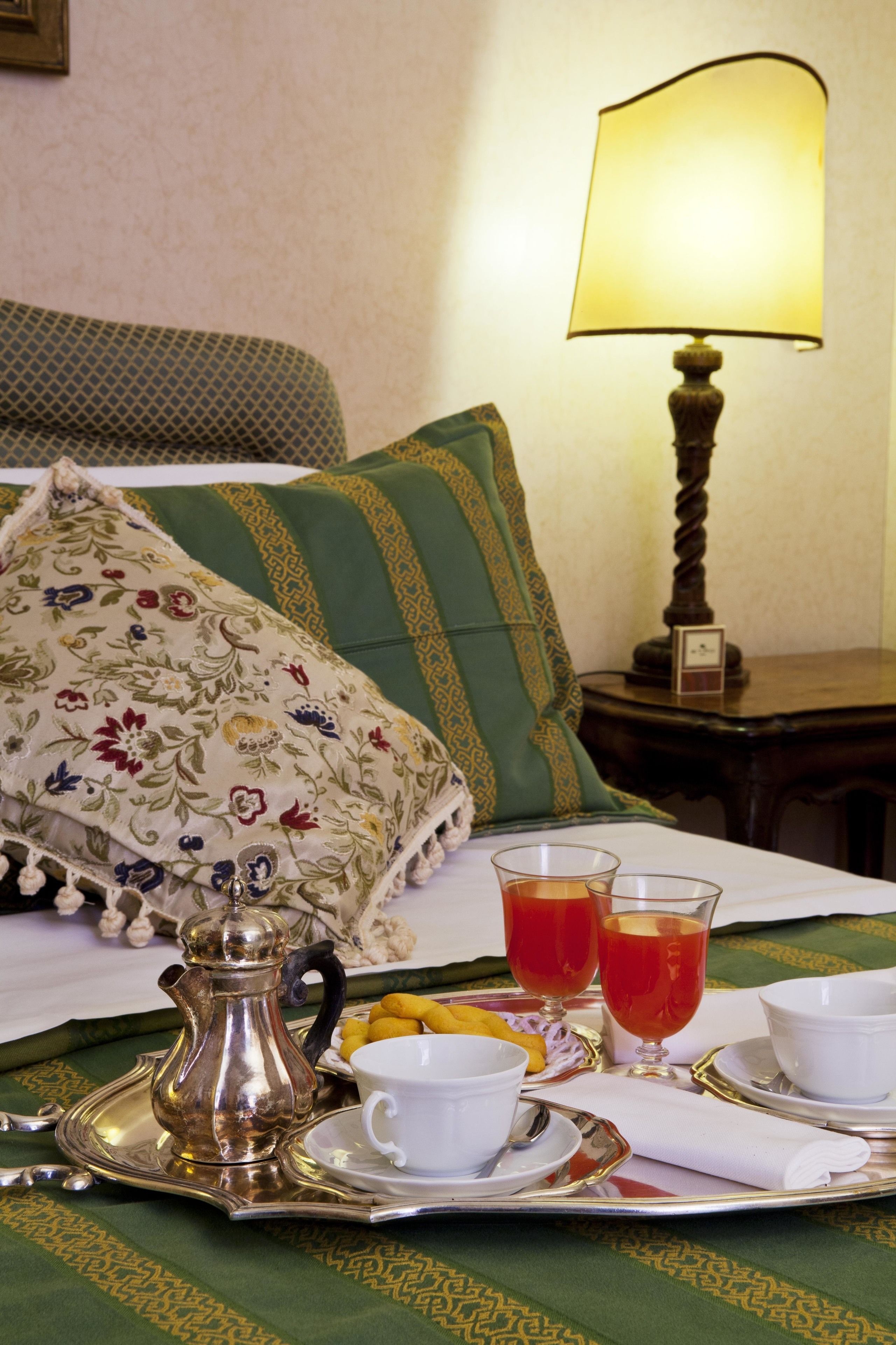 A neatly arranged breakfast tray with a silver pitcher, cups, and glasses of tea on a bed with green and floral pillows.