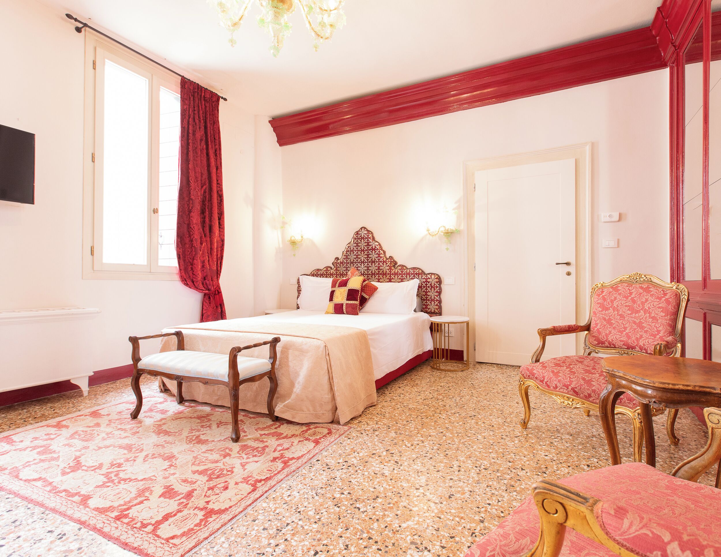 A hotel room with a large bed, a sitting area with a chair and table, a red curtain, and a chandelier.
