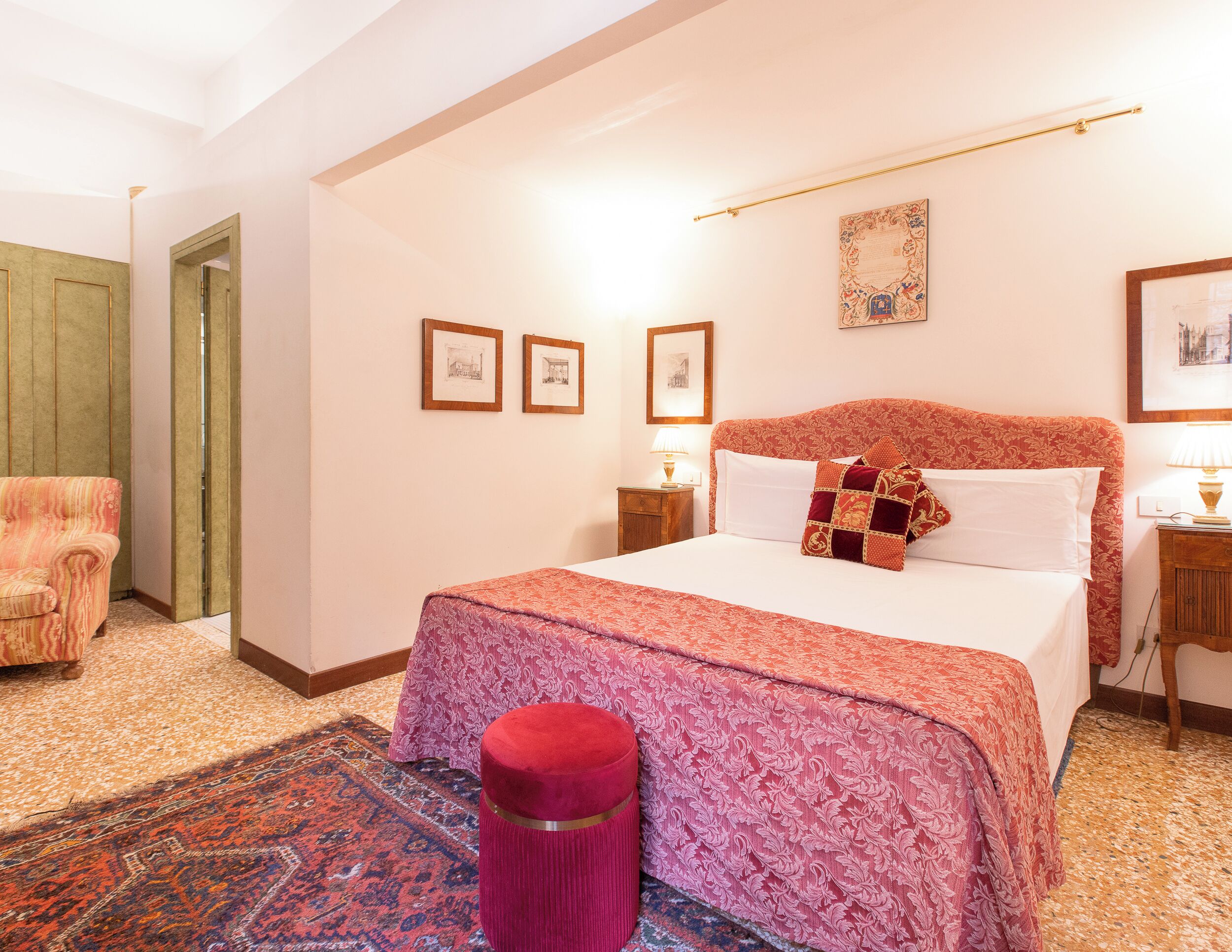 Une chambre avec un grand lit, un pouf rouge, un tapis à motifs et des tableaux encadrés accrochés au mur.