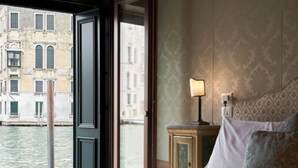 A hotel room with a bed, a nightstand with a lamp, a view of a canal, and a window with a curtain. - Palazzetto Pisani (Venice)