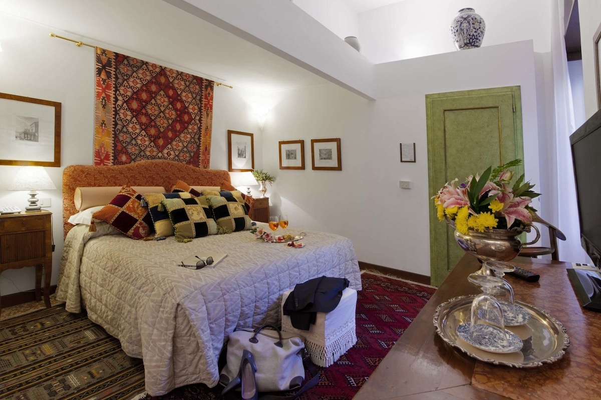 A bedroom with a bed, a nightstand, a lamp, a vase with flowers, a green door, and a patterned rug.