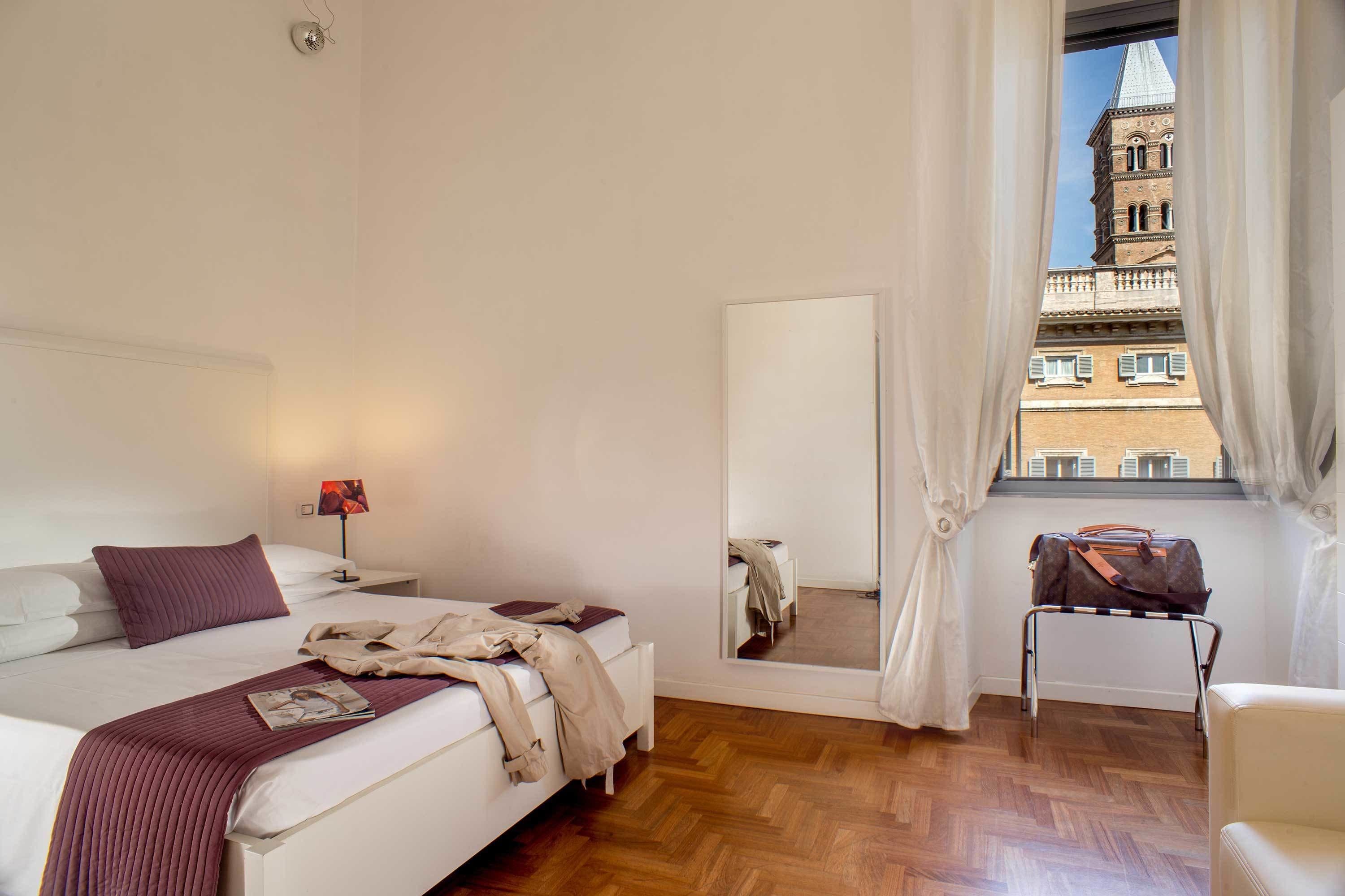 A hotel room with a bed, a bedside table with a lamp, a chair, a window with curtains, and a view of a building outside.