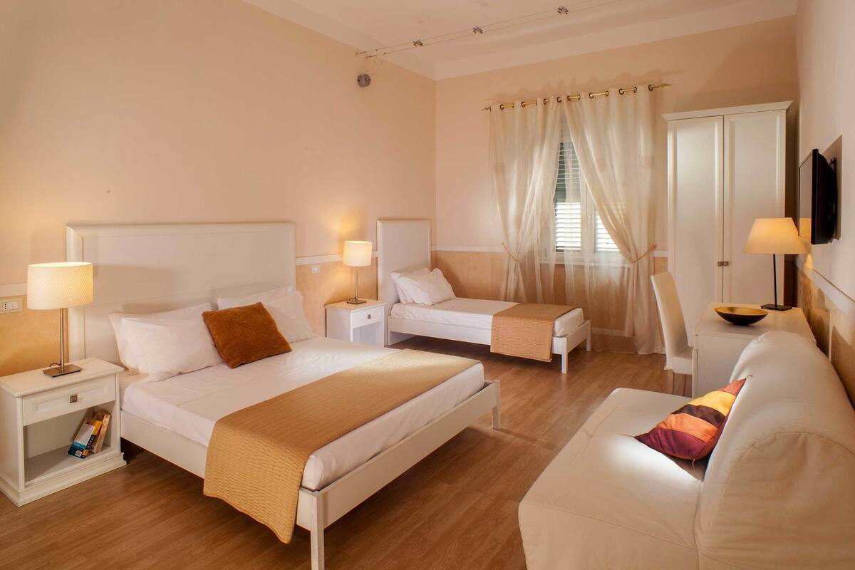 A hotel room with a large bed, two smaller beds, a sofa, a TV, and a window with curtains.