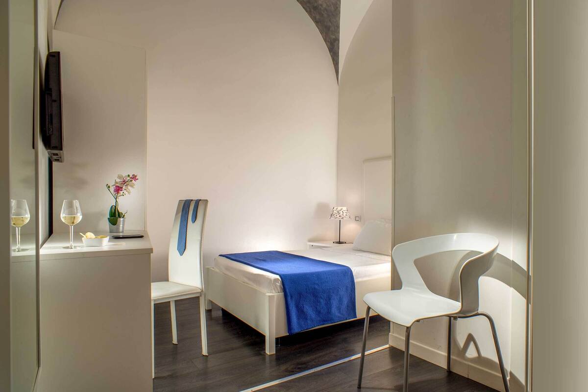 A modern hotel room with a bed, two white chairs, a desk with a laptop, a vase with flowers, and a television on the wall.