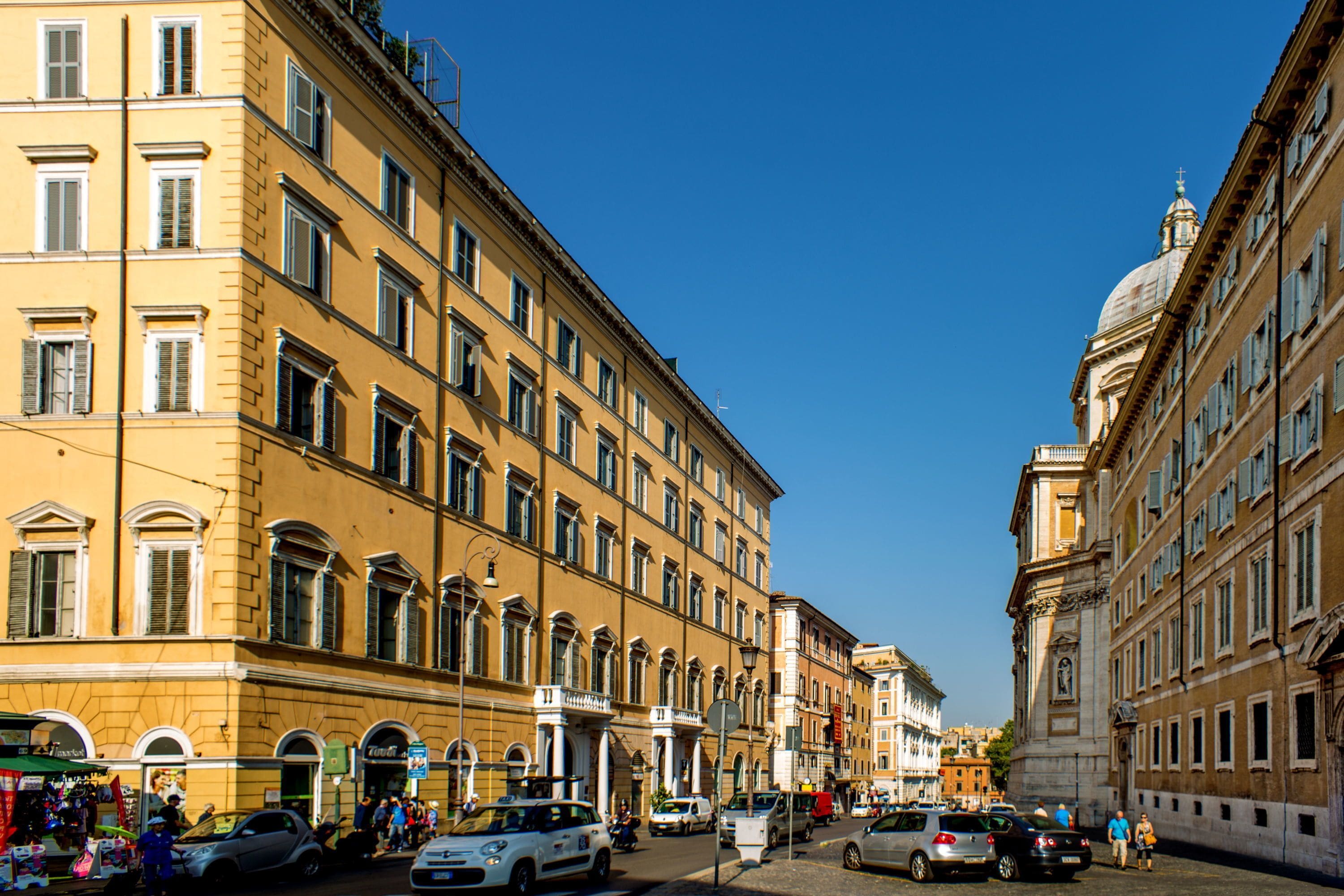 A street with historic buildings, parked cars, and pedestrians.