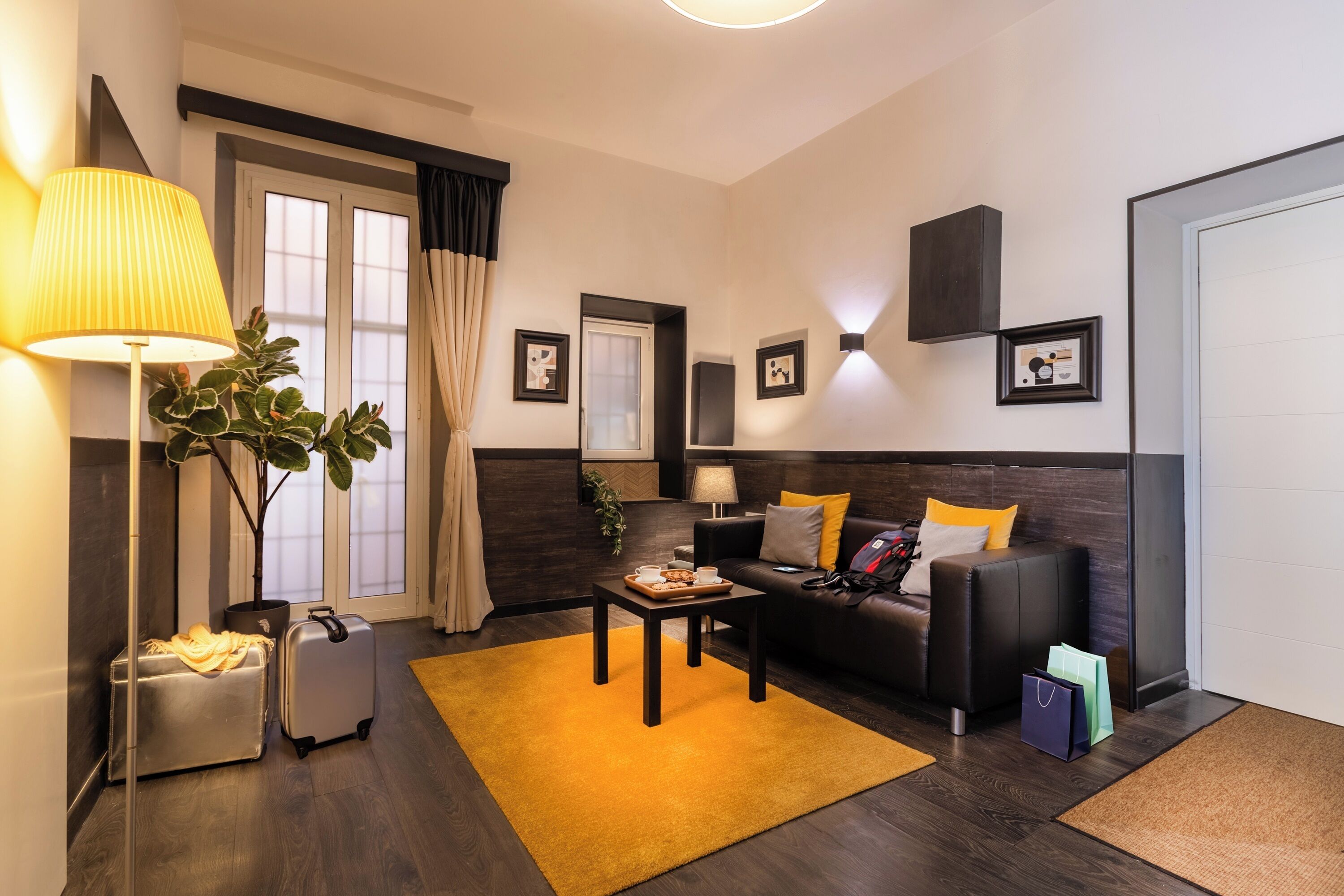 A modern living room with a sofa, coffee table, and luggage, featuring a large window, dark wood flooring, and a yellow rug.