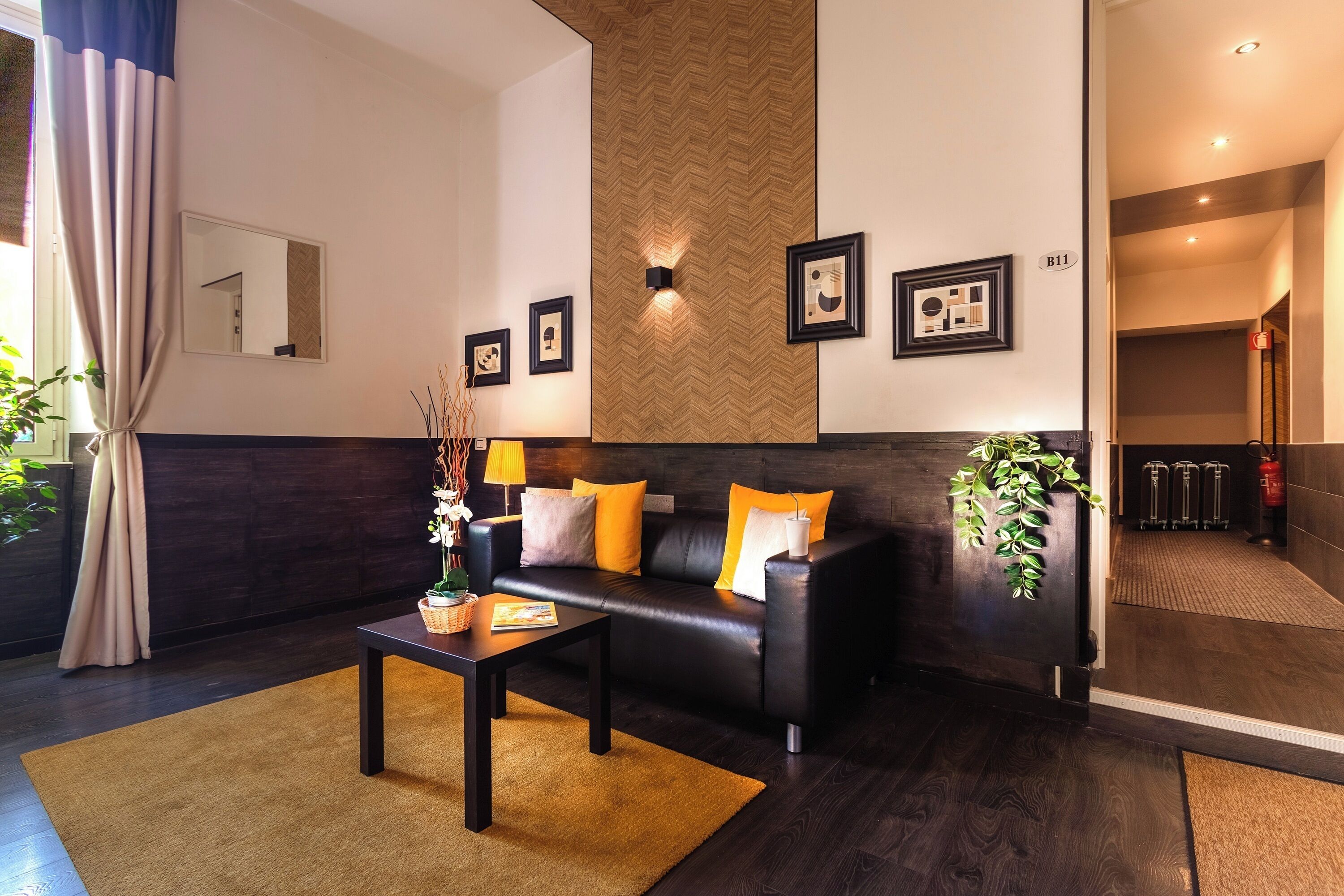 A modern hotel lobby with a dark wood floor, a black leather sofa with orange cushions, a small table with a vase of flowers, and framed pictures on the wall.
