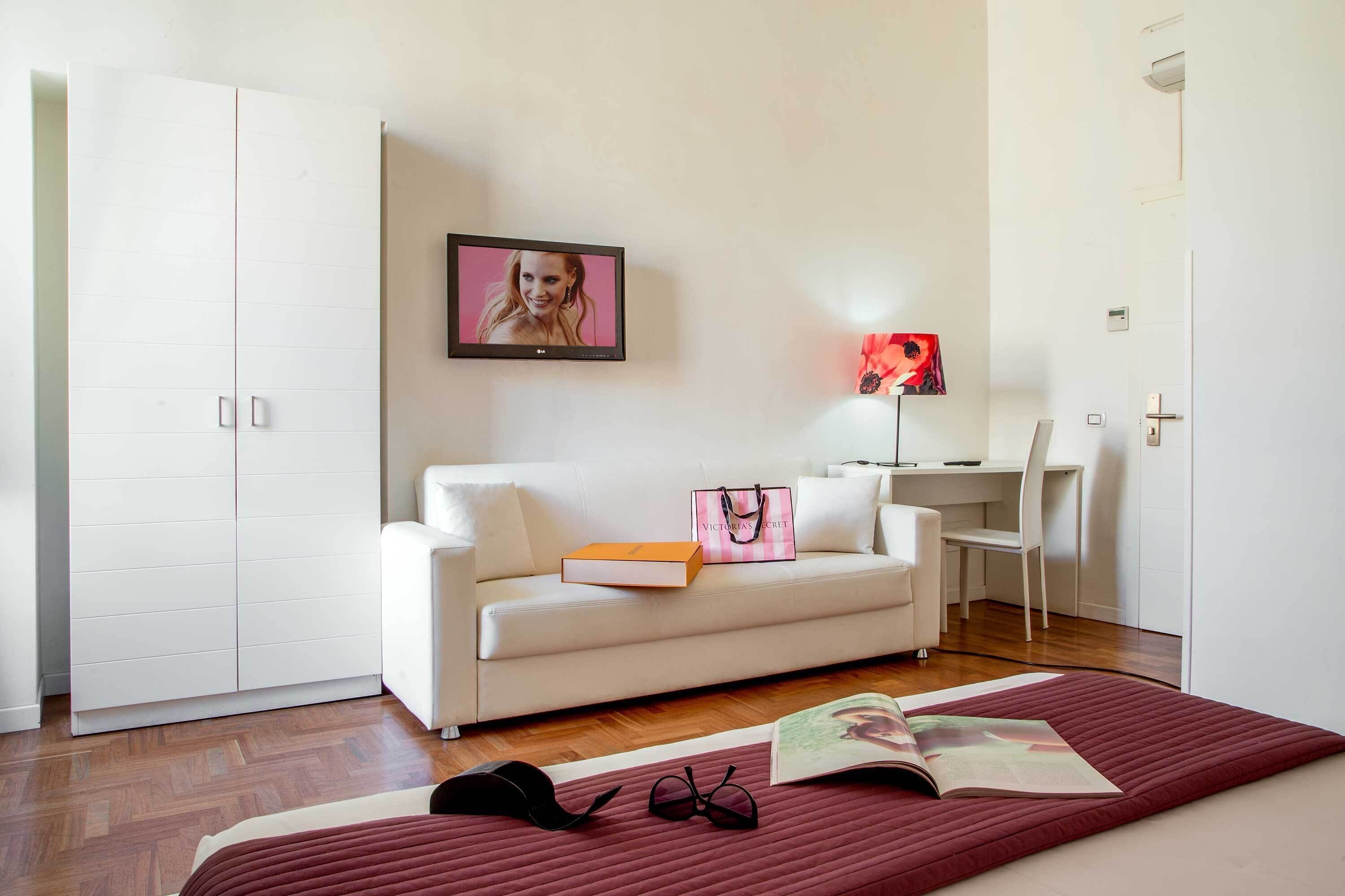 A modern living room with a white sofa, a flat-screen TV, a desk with a chair, and a red rug.