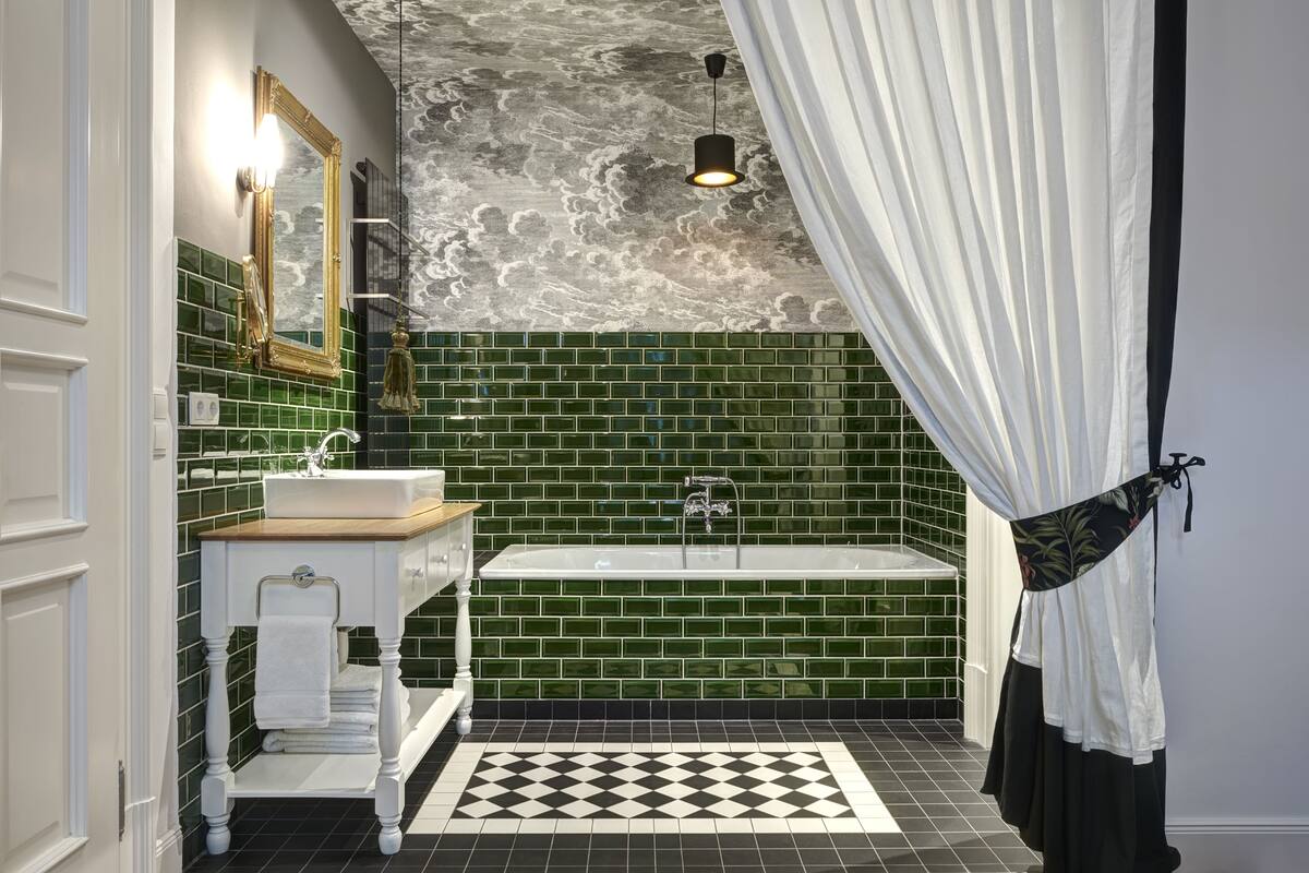 A bathroom with a green tiled wall, a white sink, a white vanity, a black and white checkered floor, a gold-framed mirror, and a black pendant light.