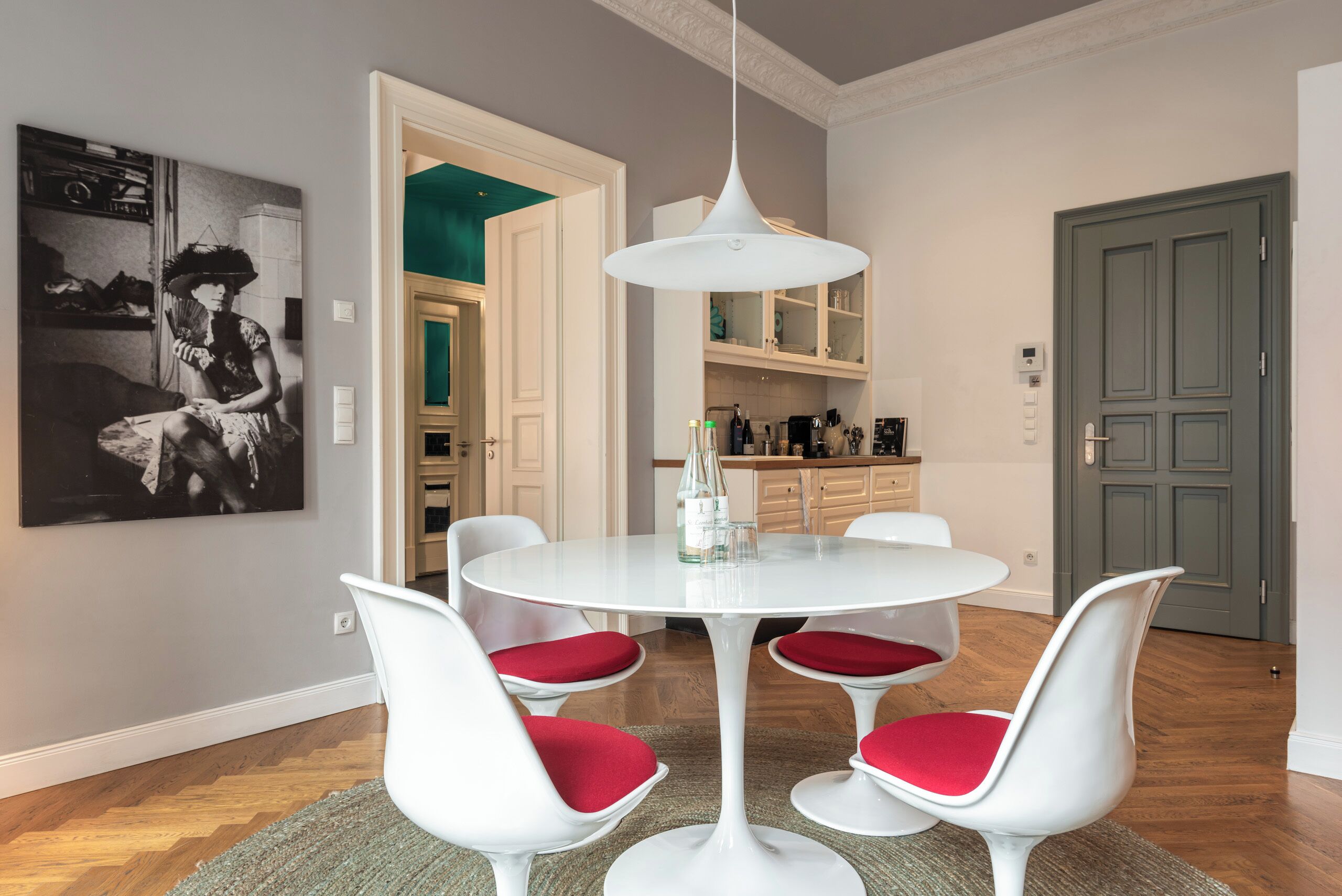 A dining area with a white round table, four white chairs with red cushions, a white pendant light, and a black and white artwork on the wall.