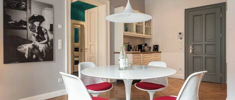 A dining area with a white round table, four white chairs with red cushions, a white pendant light, and a black and white artwork on the wall.