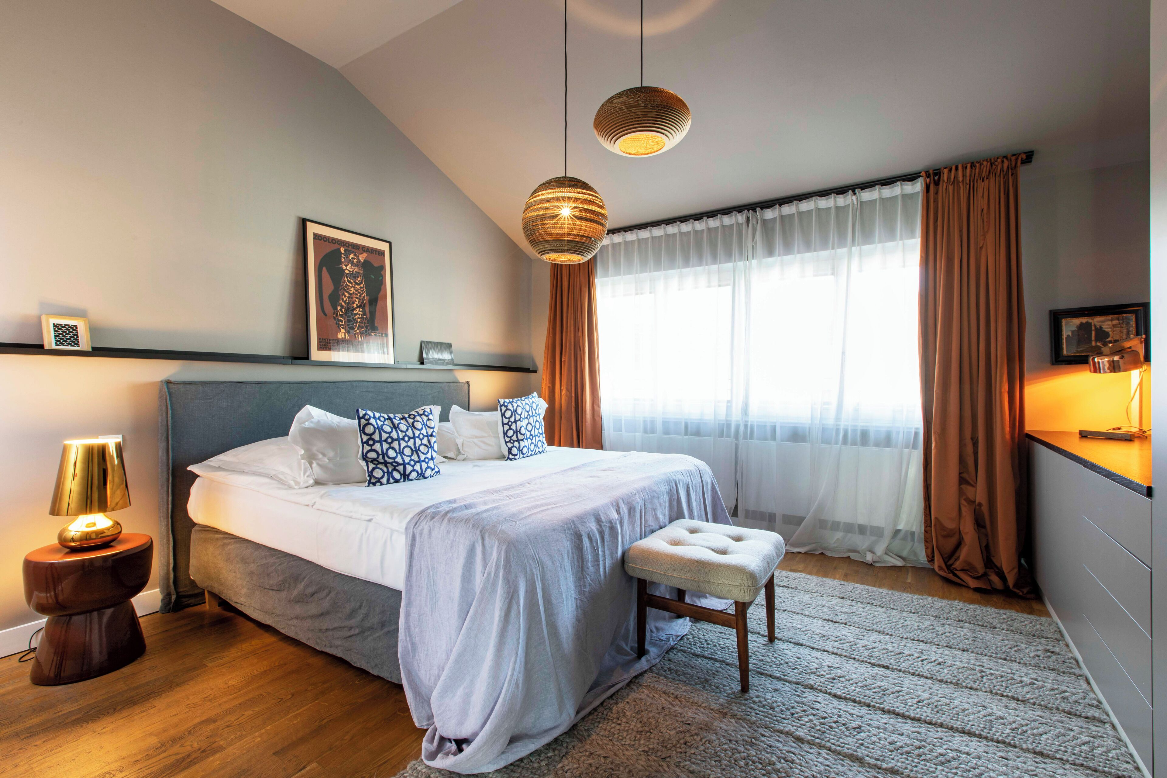 A modern bedroom with a large bed, bedside lamps, a bedside table, a bench, and a window with curtains.