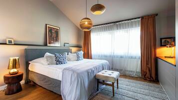 A modern bedroom with a large bed, bedside lamps, a bedside table, a bench, and a window with curtains.
