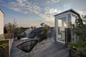 Eine Dachterrasse mit Glasumrandung, Holzdeck und schwarzen Metallstühlen.