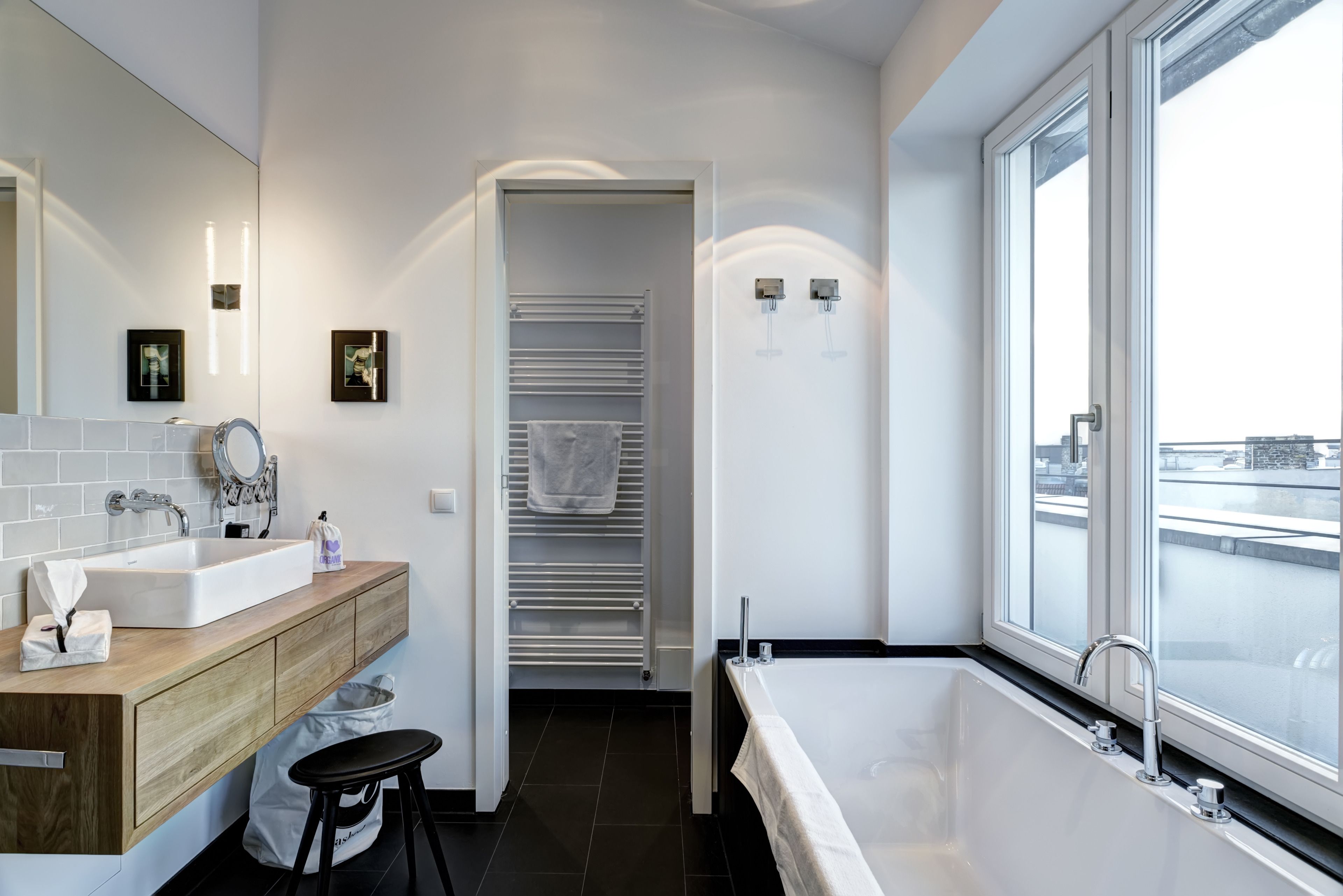 A modern bathroom with a large bathtub, a sink, and a mirror. There is a towel rack and a window with a view of the city.