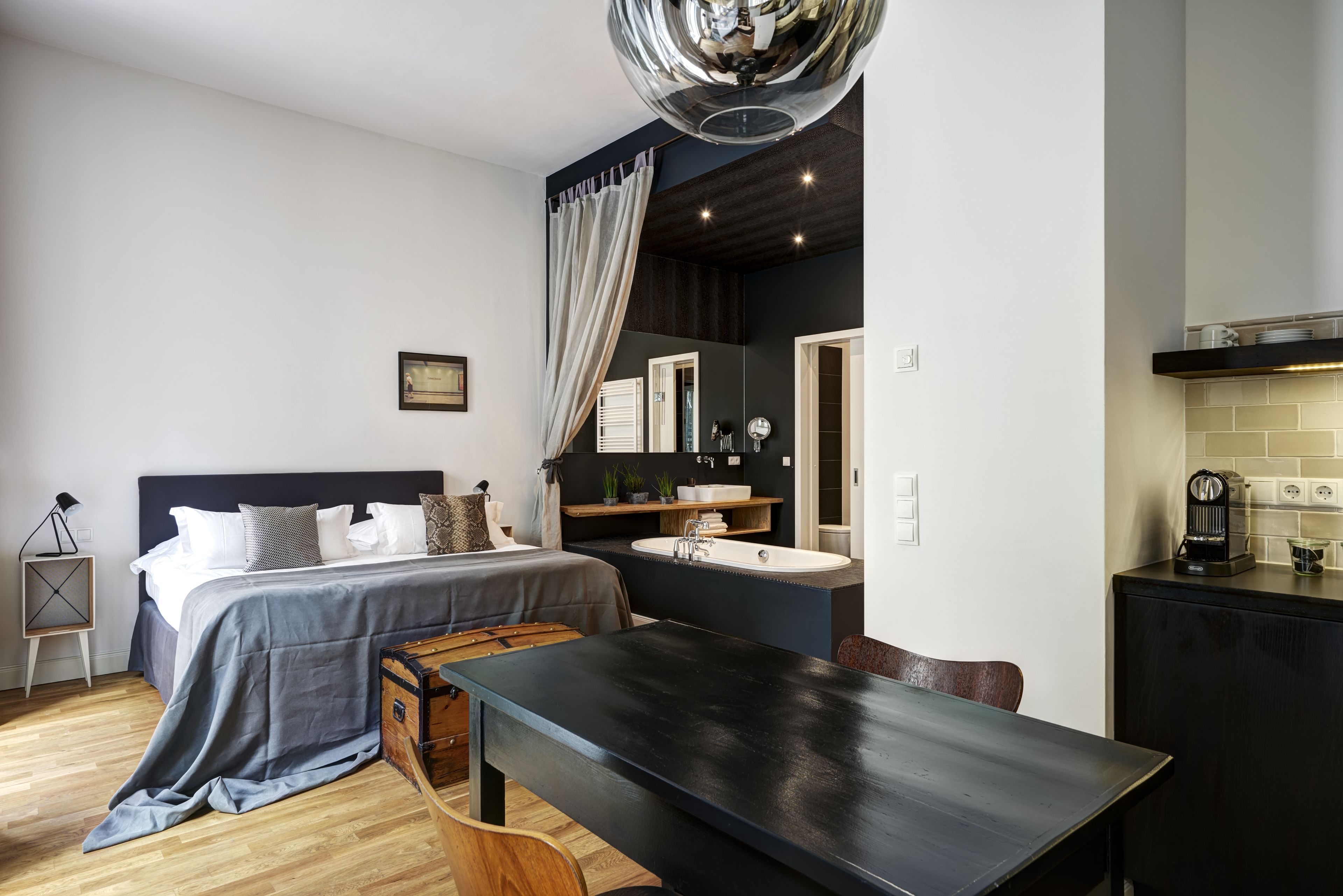 A modern bedroom with a large bed, bedside tables, a desk, and a small kitchenette.