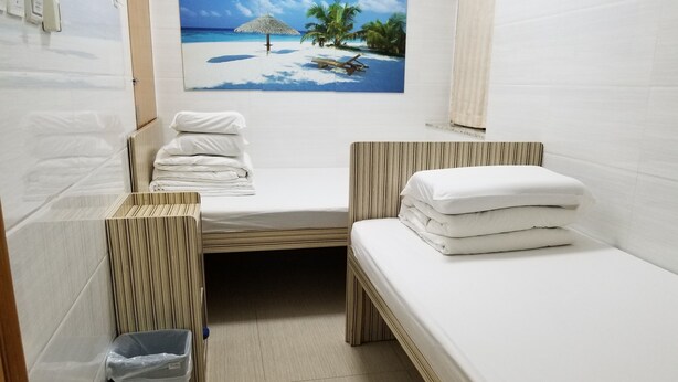Une petite pièce propre, avec deux lits, une poubelle et un tableau représentant une scène de plage accroché au mur.