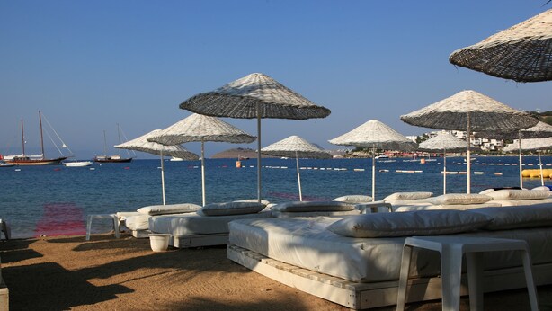 A beachside lounge area with white lounge chairs and thatched umbrellas, overlooking a calm sea with boats. - Okaliptus Hotel (Bodrum)