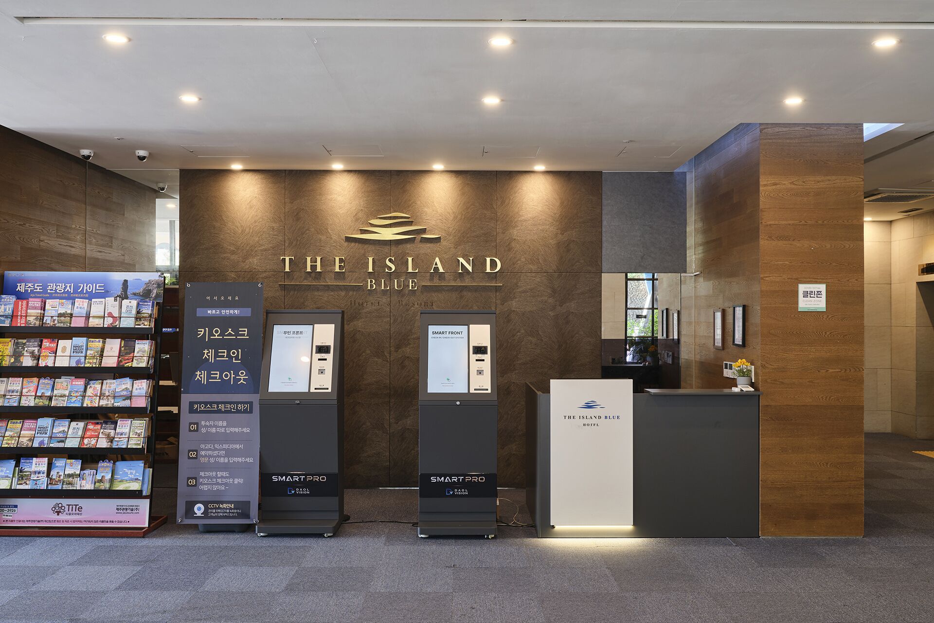 Una zona d'arribada amb un cartell que diu "THE ISLAND BLUE HOTEL". Hi ha dues quioscs d'autoatenció etiquetats "SMART PRO". Es veu una biblioteca amb fulletons turístics a l'esquerra.