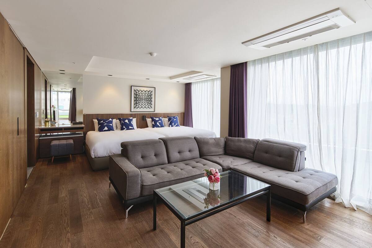 A modern hotel room with a large bed, a grey sectional sofa, a glass coffee table, and a view of the city through large windows.