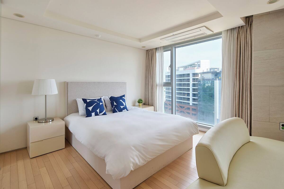 A modern hotel room with a large bed, a bedside lamp, a chair, and a view of the city through large windows.