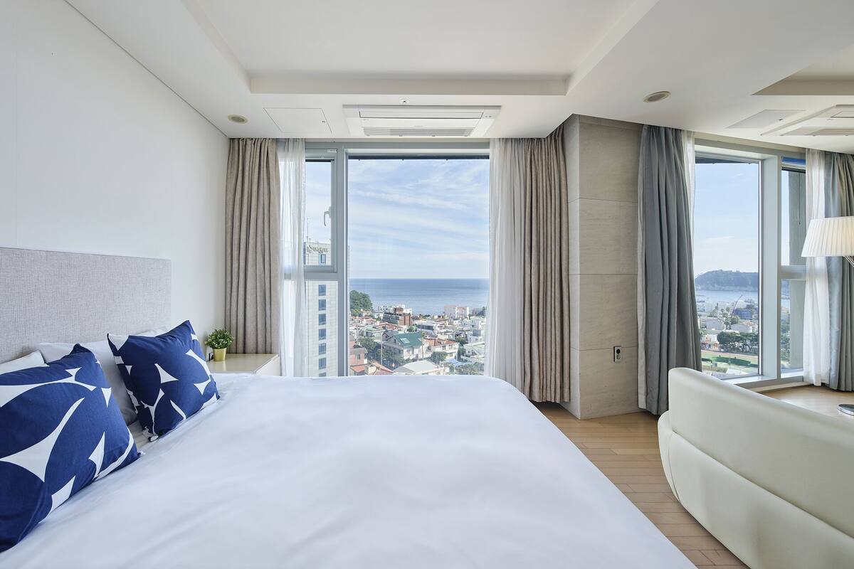 A modern hotel room with a large bed, a sofa, and a view of the city and sea.