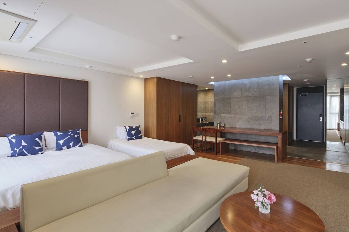 A modern hotel room with two beds, a sofa, a dining area, and a bathroom visible through an open door.