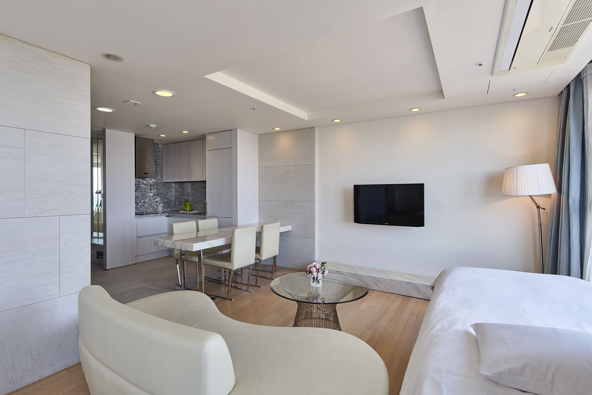 A modern hotel room with a bed, a seating area with a glass table, a kitchen area, and a TV mounted on the wall.
