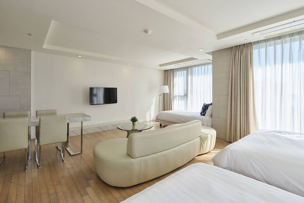A modern hotel room with a large bed, a sofa, a dining area with chairs, a TV mounted on the wall, and a window with curtains.
