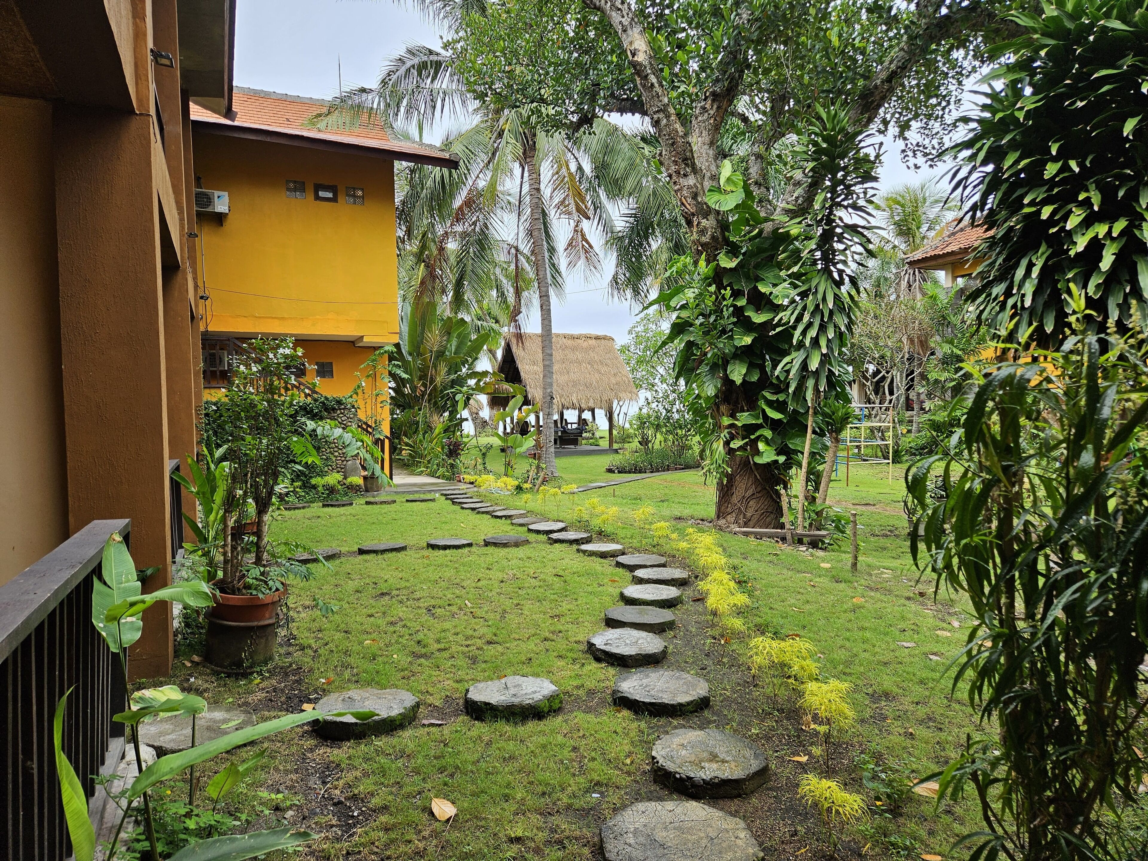 A pathway with stepping stones leading to a thatched-roof structure surrounded by lush greenery and a yellow building.